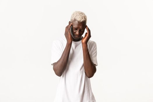 Portrait of dizzy african-american blond man, feeling headache, grimacing and bending from pain, having migraine, standing white background.