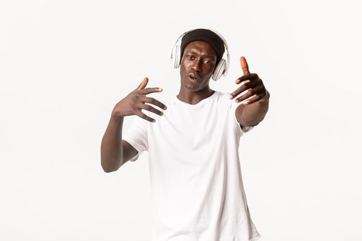 Portrait of joyful cool african-american guy, dancing and listening music in headphones, white background.