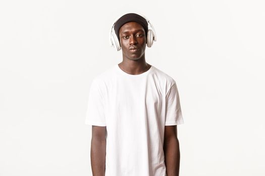 Portrait of cool handsome african-ameircan guy in beanie, listening music in wireless headphones, looking serious, white background.