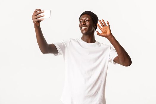 Portrait of happy smiling african-american guy talking with friends via video call, holding smartphone and waving hand in hello gesture, standing white background.