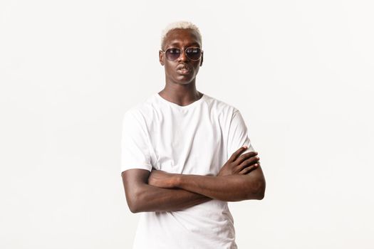 Portrait of confident and serious african-american blond guy wearing sunglasses and cross arms chest, acting sassy and bossy, standing white background.