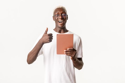 Portrait of happy and satisfied, handsome african-american blond male, wearing glasses, holding digital tablet and showing thumbs-up, reading social network or books, white background.