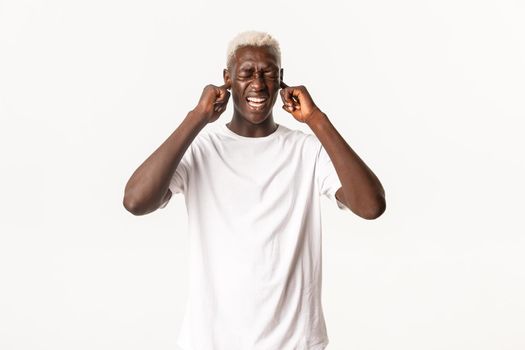 Portrait of annoyed african-american blond man, shut ears with fingers and grimacing bothered, hate loud awful noise, white background.