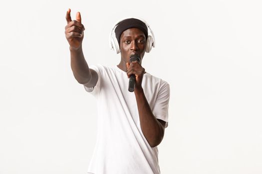 Portrait of sassy and cool african-american guy in wireless headphones, singing into microphone, standing white background.