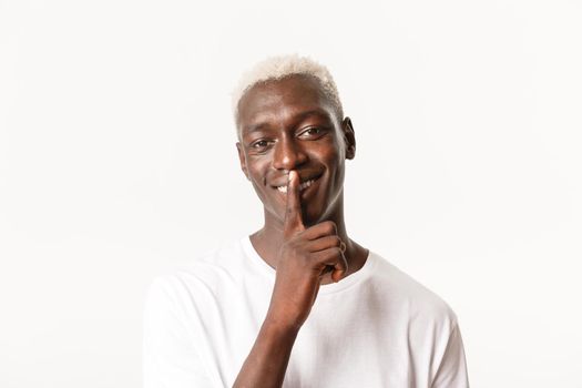 Portrait of happy attractive Black guy with blond hair, showing shush gesture and smiling, prepare surprise, need silence, white background.