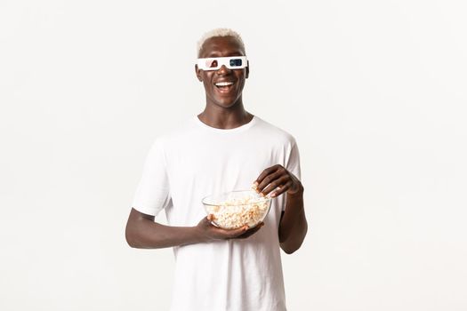 Portrait of carefree smiling african-american guy, watching movie in 3d glasses and eating popcorn, enjoying cinema, standing white background.