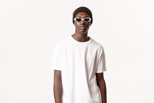 Portrait of stylish and cool african-american young guy in beanie and sunglasses, looking confident and sassy, standing over white background.