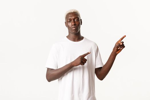 Portrait of serious african-american blond guy, pointing fingers upper right corner and showing logo, standing white background.