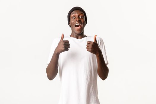 Portrait of excited, cheerful african-american guy in beanie, looking amused and showing thumbs-up in approval, white background.