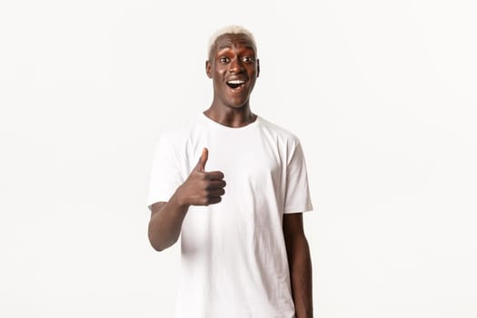 Portrait of impressed smiling african-american blond man, showing thumbs-up, praising good choice, making compliment, white background.