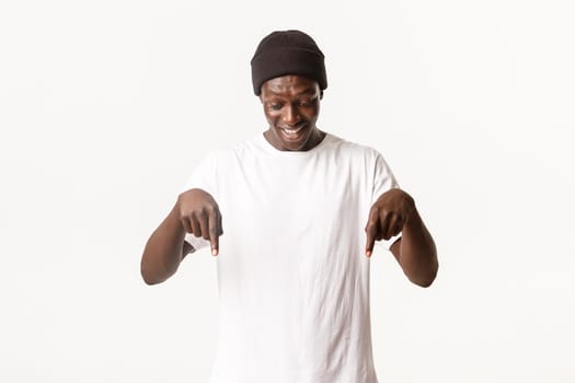 Portrait of happy and amused african-american handsome guy in beanie, looking and pointing fingers down at something interesting, white background.