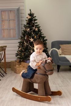 Portrait of little adorable toddler sits on a brown textile rocking horse, in the living room, with a Christmas tree, looks at to camera. Vertical