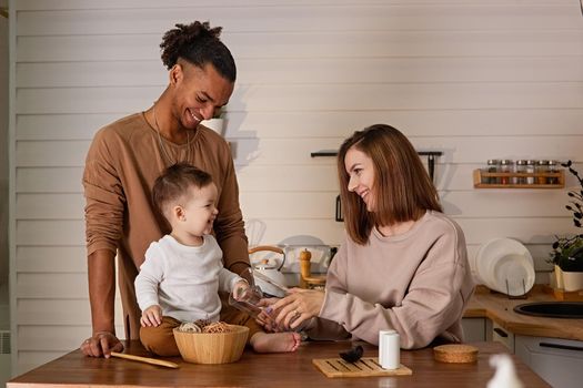 Happy young multiracial family, in cozy brown clothes, sitting at the table in the kitchen, having fun with their little toddler son