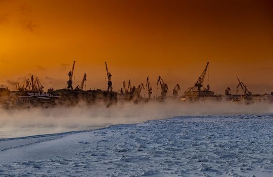 The construction of nuclear icebreakers at magic sunset, cranes of of the Baltic shipyard in a frosty winter day, steam over the Neva river, smooth surface of the river, sky of orange color. High quality photo