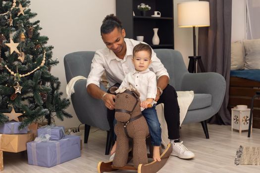 A young stylish father sits on a gray sofa, rolls his little toddler son on a brown rocking horse, at home, in the evening.