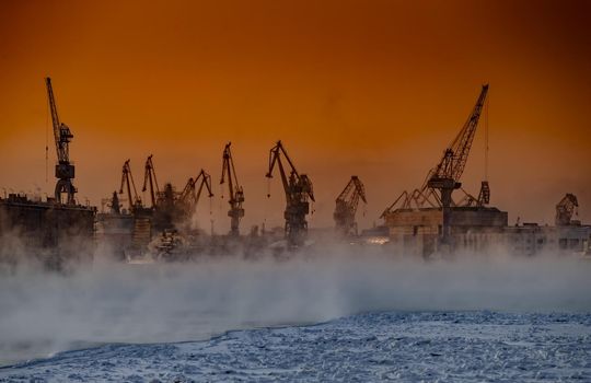 The construction of nuclear icebreakers at magic sunset, cranes of of the Baltic shipyard in a frosty winter day, steam over the Neva river, smooth surface of the river, sky of orange color. High quality photo