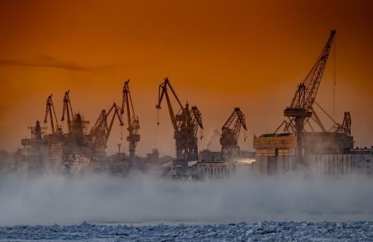 The construction of nuclear icebreakers at magic sunset, cranes of of the Baltic shipyard in a frosty winter day, steam over the Neva river, smooth surface of the river, sky of orange color. High quality photo