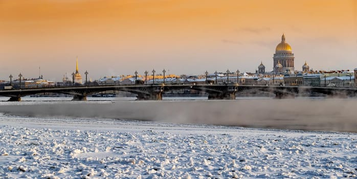 Winter panoramic view of St. Petersburg at sunset, Isaac cathedral and Blagoveshenskiy bridge on background, steam over frozen Neva river, sky of orange color. High quality photo