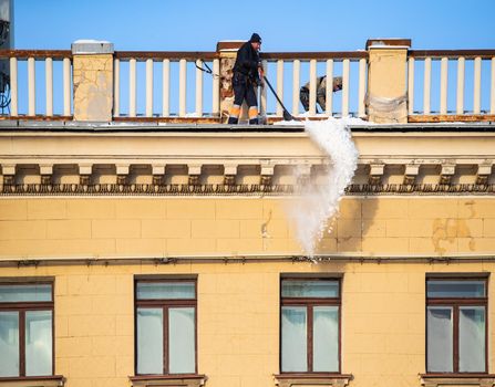 Russia, St. Petersburg, 07 December 2021: The work of utilities to remove snow from the roofs of houses, a man throws off the snow of the roof, he is attached with insurance to the roof structure. High quality photo