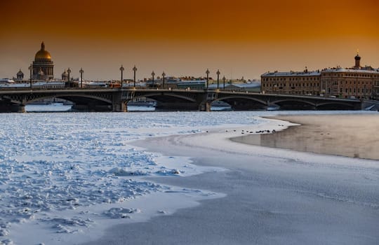 Winter panoramic view of St. Petersburg at sunset, Isaac cathedral and Blagoveshenskiy bridge on background, steam over frozen Neva river, sky of orange color. High quality photo