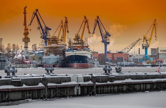 The construction of nuclear icebreakers at magic sunset, cranes of of the Baltic shipyard in a frosty winter day, steam over the Neva river, smooth surface of the river, sky of orange color. High quality photo