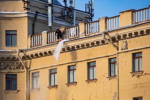 Russia, St. Petersburg, 07 December 2021: The work of utilities to remove snow from the roofs of houses, a man throws off the snow of the roof, he is attached with insurance to the roof structure. High quality photo