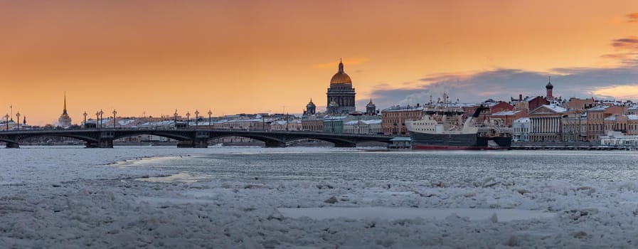 Winter panoramic view of St. Petersburg at sunset, Isaac cathedral and Blagoveshenskiy bridge on background, steam over frozen Neva river, huge ship moored near embankment, sky of orange color. High quality photo