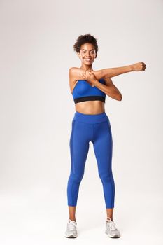 Full length of beautiful smiling african-american sportswoman, wearing blue sport outfit, warming-up before workout, showing stretching exercises, white background.