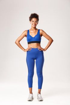 Full length of confident athletic african-american girl in sportswear, smiling pleased, holding hands on waist with determined expression, workout over white background.