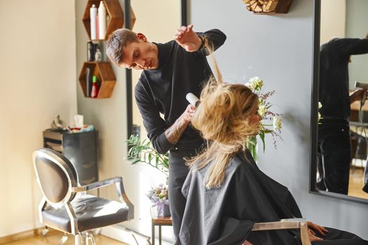 Happy beautiful light hair caucasian woman at beauty salon. Male master is drying her hair