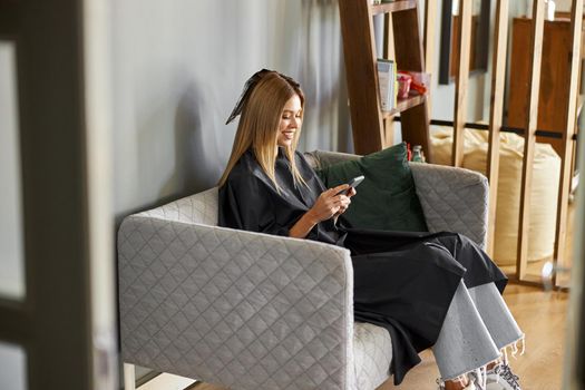 Happy beautiful light hair caucasian woman at beauty salon is sitting on sofa and using smartphone