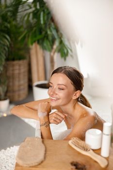 Joyful young woman smiling away and leaning on bathtub side while bathing at spa resort. Wellness, beauty and care concept