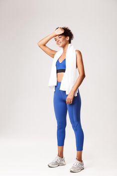Full length of satisfied smiling, fit woman in blue sportswear, standing over white background with towel, wiping sweat off forehead after workout.