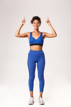 Full length of attractive slim african-american sportswoman, athlete pointing fingers up at logo or brand advertisement, standing in workout outfit over white background.