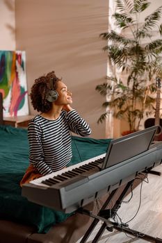 Multiracial woman sitting on bed and smiling while playing melody on synth electronic musical instrument