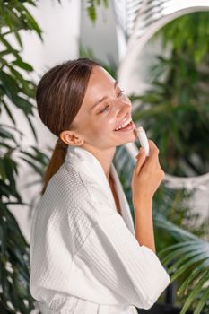 Portrait of cute young woman with beautiful skin smiling while applying lip balm on her lips, standing in the bathroom. Skincare cosmetics concept