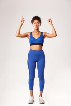 Full length of amazed good-looking fitness girl in blue sports bra and leggings, pointing fingers up, showing promo about workout or gym, standing over white background.