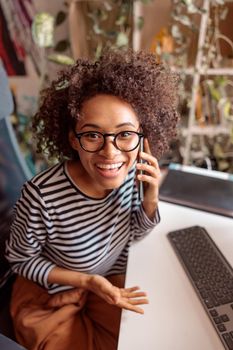 Cheerful multiethnic lady having phone conversation and smiling while sitting at the table with keyboard