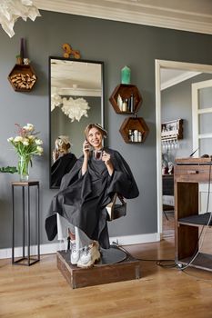 Happy blonde caucasian woman is using smartphone while sitting in chair and waiting in hairdresser's salon
