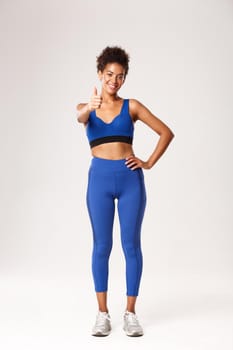Full length of attractive african american sportswoman in fitness clothing, showing thumbs-up in approval, recommend sport brand, standing over white background.