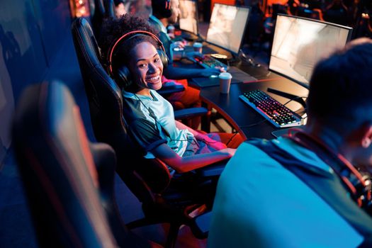 African female cybersport gamer wearing headset sitting on gaming chair and looking at camera with cheerful expression in cyber club