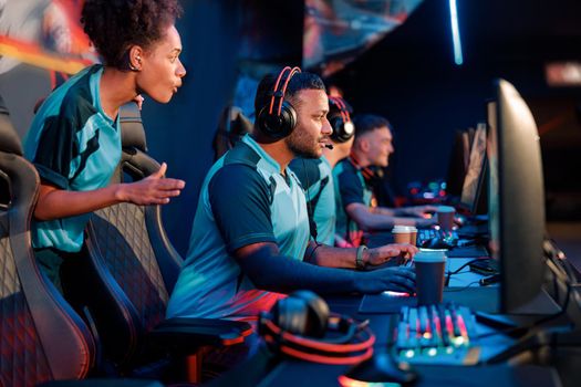 Young African female, cybersport gamer cheers male friend while participating as one team in esports tournament in gaming club