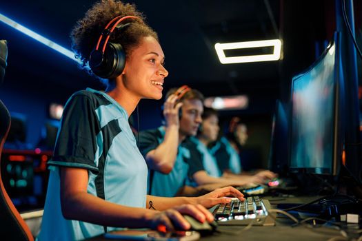 Happy African female cybersport gamer wearing headphones playing online video game while participating in esports tournament in internet cafe