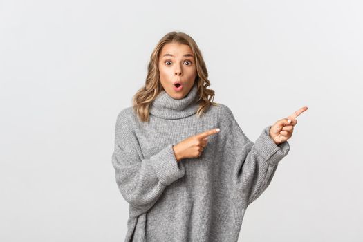 Amazed and excited blond girl in grey sweater showing banner, pointing fingers right and looking at camera, standing over white background.