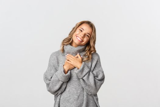 Close-up of tender blond girl in grey sweater, holding hands on heart and looking touched and thankful, smiling as appreciate something, standing over white background.