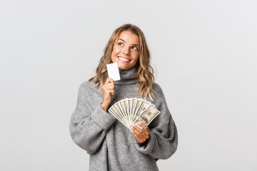 Beautiful blond girl looking thoughtful and smiling, holding credit card and money, standing over white background.