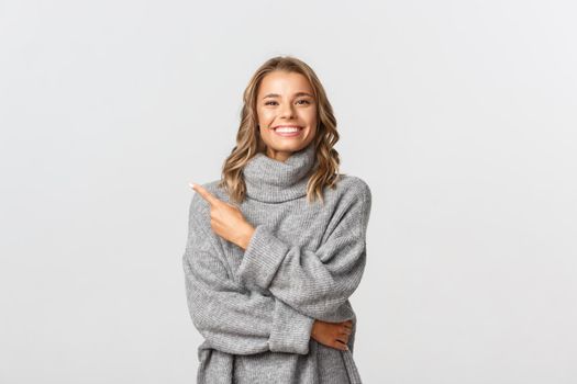 Image of beautiful blond girl in grey sweater pointing finger at upper left corner, showing your logo or banner, standing over white background.