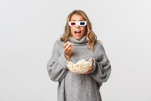 Image of thrilled blond girl in grey sweater and 3d glasses, holding bowl with popcorn, and gasping amazed while watching movie, standing over white background.