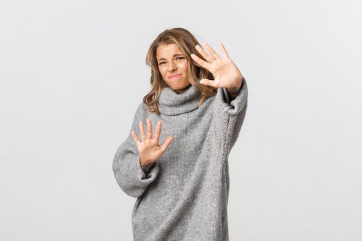 Image of disturbed blond woman feeling uncomfortable and asking to stop photographing, stretching hands forward to cover face from light, standing over white background.