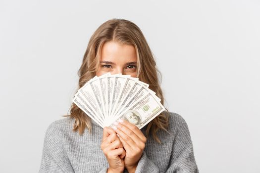 Close-up of excited pretty girl in grey sweater, peeking through money, thinking about shopping, standing over white background.